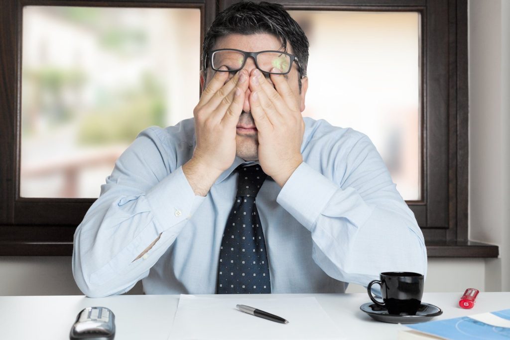 Stressed man at desk in bright office, struggling with overwhelming workload and frustration.