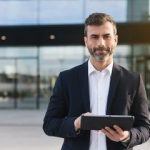 Confident businessman in tailored suit holding tablet in front of modern glass building.