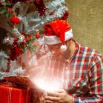 a man in a santa claus hat opening a gift under a christmas tree