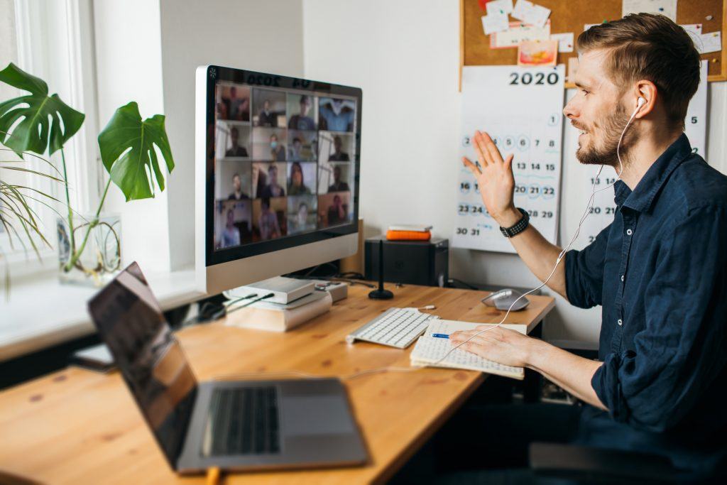 man engaged in a virtual meeting at his home office desk with laptop and monitor.