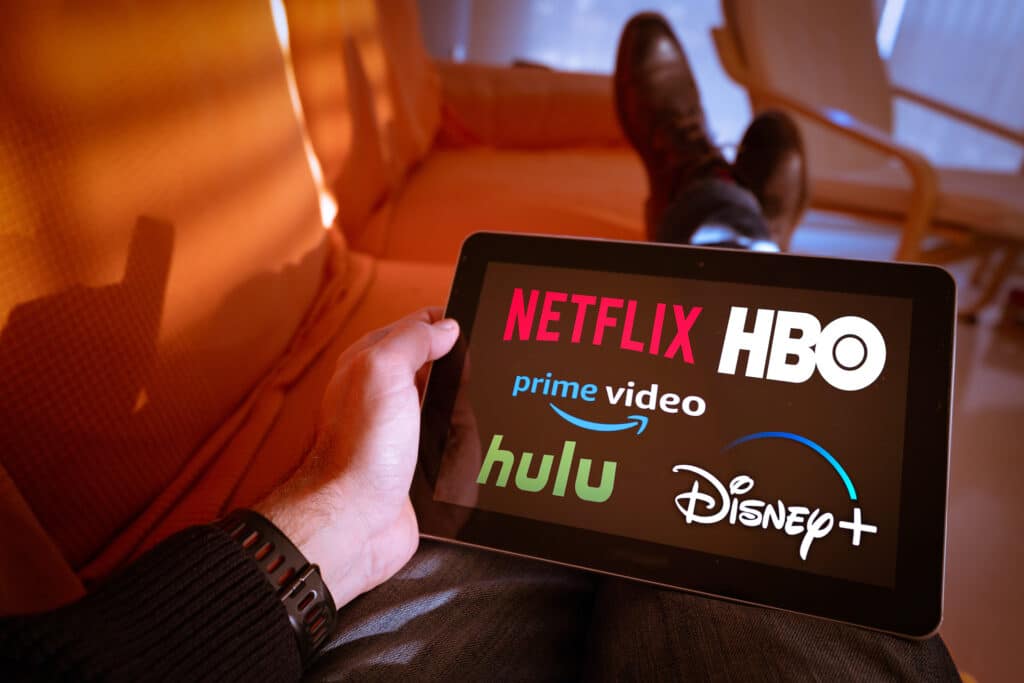 man relaxing with tablet showing logos of streaming services netflix, hulu, hbo, amazon, and disney+.