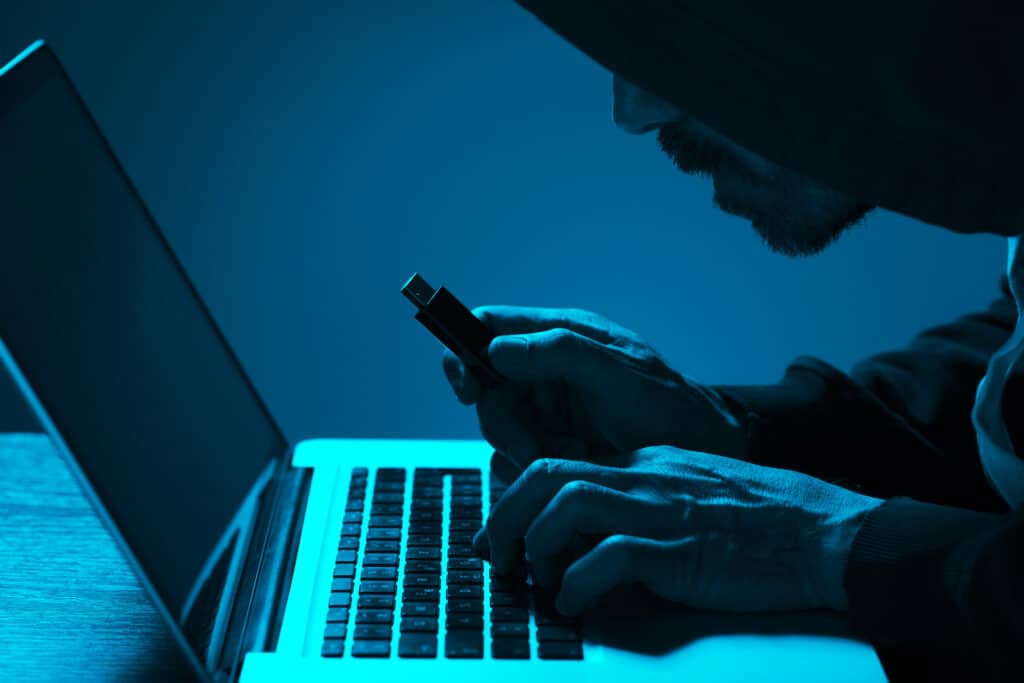 hooded hacker stealing data from a laptop in a dimly lit room with usb drive.